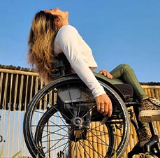 Foto de mulher branca, de cabelos claros e longos, cadeirante, em um local de grama. Pose olhando para o céu.